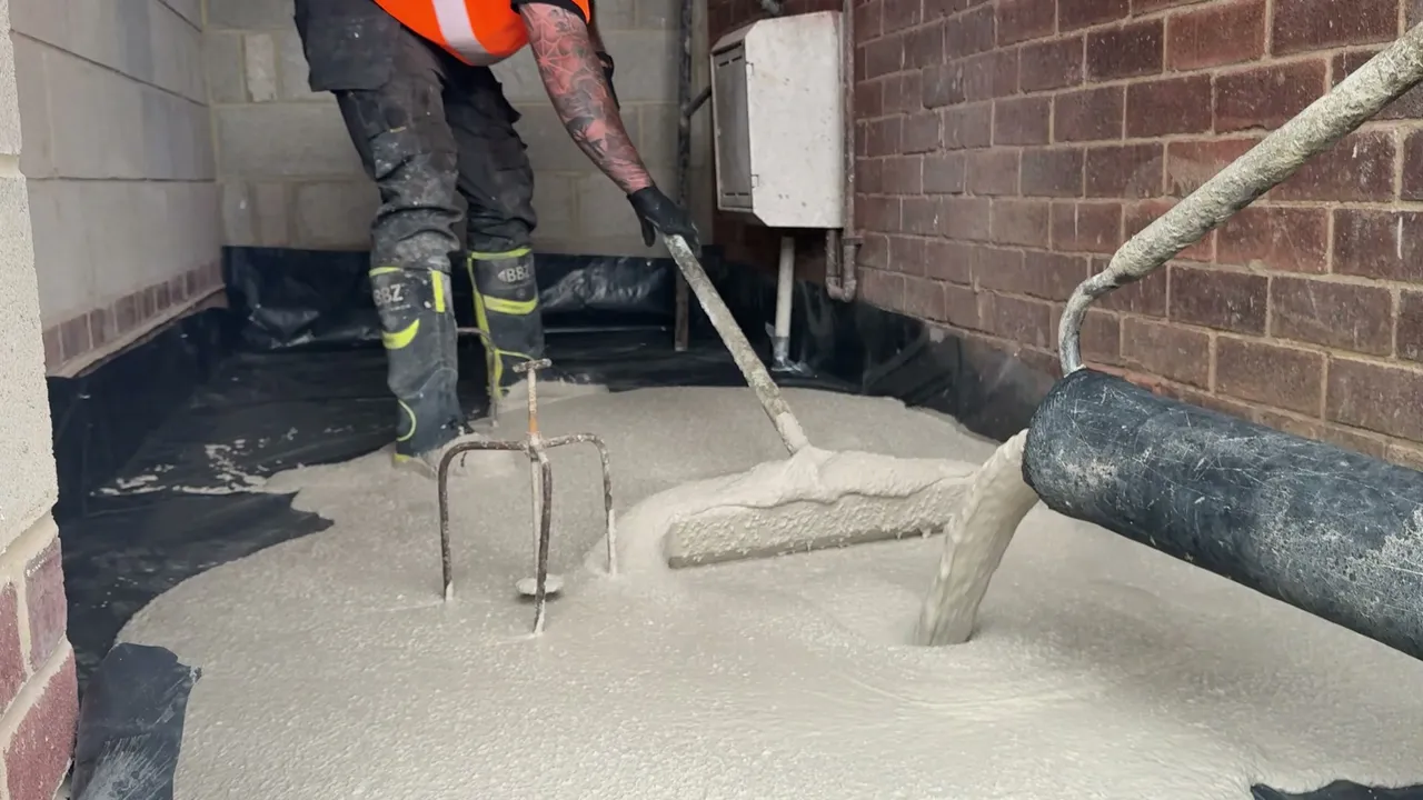 Gypsol Classic liquid screed discharging from the pump line into a room while an operative works it out with a dapple bar